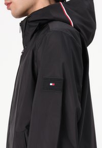 Sort letvægtsjakke med hætte. Har røde og hvide striber på hætten, lynlåslukning og et rektangulært Tommy Hilfiger-logo på ærmet.
