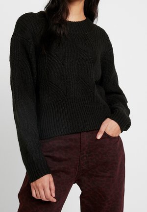 Mujer con un suéter negro de punto trenzado y pantalones rojo oscuro con un sutil estampado de leopardo, posando con una mano en el bolsillo.