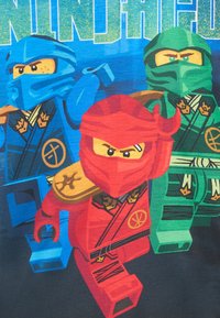 T-Shirt mit drei LEGO-Ninja-Charakteren: rot, blau und grün. Jeder trägt einzigartige Masken und Rüstungen mit komplexen Mustern, vor einem dunklen Hintergrund.