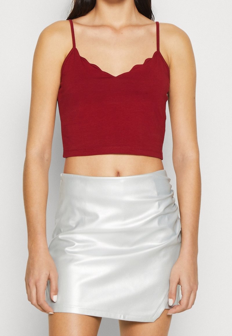 Femme portant un crop top rouge à bords festonnés avec de fines bretelles et une mini-jupe argentée brillante avec un léger froncé sur un côté.