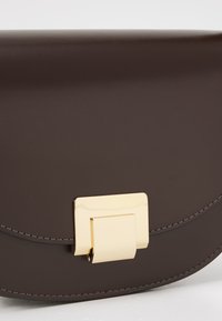 Sac à main en cuir marron foncé avec un design incurvé, doté d'un fermoir doré et d'une couture tonale le long des bords. Texture lisse.