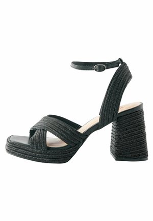 Black platform sandals feature braided straps and a wide, textured block heel. An ankle strap with a circular buckle is included.