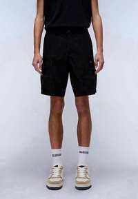 Bermudas pretas com múltiplos bolsos, corte reto, tecido durável, combinadas com ténis bege e meias brancas. T-shirt preta simples.