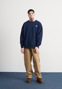 Sudadera azul marino con un logo blanco, combinada con pantalones cargo caqui y botas marrones. Silueta clásica, de corte relajado, con puños acanalados.