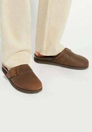 Brown leather slip-on shoes with a rounded toe, featuring a side strap and buckle detail, paired with light-colored trousers.