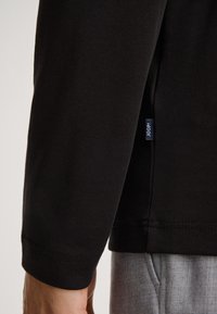 Schwarzes Langarmshirt aus weicher Baumwolle. Besitzt eine subtile Textur, enganliegende Manschetten und ein kleines Markenlabel an der Seitennaht.