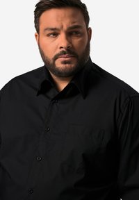 Musta nappikauluspaita, jossa on terävä kaulus ja yksi rintatasku. Silkkinen pinta, jossa on hienovarainen kiilto, mustat napit ja vartalonmyötäinen leikkaus.