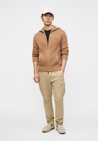 Πλεκτό ζακέτα με φερμουάρ σε ταμπά χρώμα, με μαύρο μπλουζάκι από κάτω, συνδυασμένο με ταμπά παντελόνια cargo και ανοιχτό γκρι παπούτσια χωρίς κορδόνια. Λευκό καπέλο ως τόνωση.