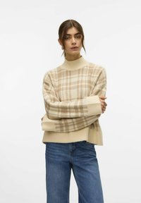 Beżowy sweter typu golf z prążkowanym wzorem w jasnobrązową kratkę. Noszony na niebieskich dżinsach, charakteryzujący się luźnym krojem.