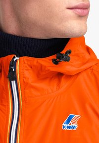 Veste légère orange vif avec un col montant, textile côtelé bleu marine contrastant, fermeture éclair à l'avant et logo K-Way sur la poitrine.