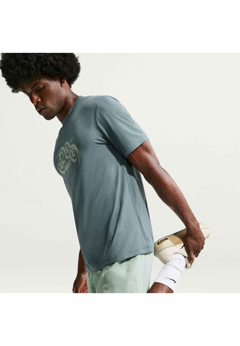 Homme en tenue de sport étirant le quadriceps en tenant un pied derrière lui, portant un t-shirt bleu et un short vert clair.