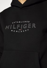 Felpa nera con cappuccio realizzata in morbido tessuto, caratterizzata da una grafica bianca che riporta la scritta "ESTABLISHED HILFIGER MCMXLXV" sulla parte frontale.
