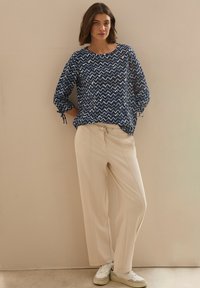 Blusa blu navy con motivo zigzag e maniche a tre quarti, abbinata a pantaloni beige chiaro a vestibilità ampia e sneakers bianche.