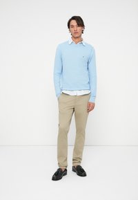 Bărbat în picioare, cu mâinile în buzunare, purtând un pulover bleu deschis peste o cămașă albă cu guler, pantaloni bej și mocasini negri cu franjuri, pe un fundal alb.