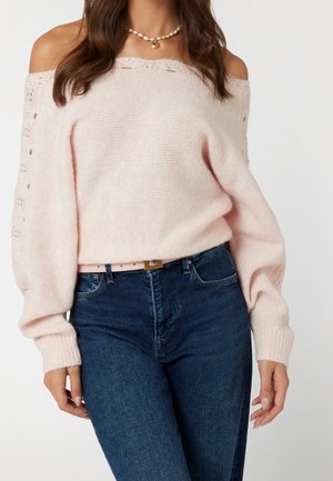 Vrouw draagt een zachtroze off-shoulder gebreide trui met kanten details en een blauwe hooggetailleerde spijkerbroek, geaccentueerd met een parelketting en een roze riem.