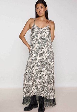 Vrouw in een zwart-witte maxi-jurk met bloemenprint, dunne bandjes, zwarte kantzoom en zwarte chunky laarzen, poserend met één hand op haar nek.