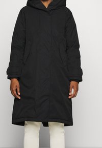 Černá parka s kapucí, která má štíhlý design, texturovanou tkaninu a boční kapsy. Obsahuje dlouhé rukávy a střih do výšky poloviny stehna.