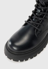 Bota de cuero negra con punta redondeada, suela de goma gruesa y un cordón intrincado. Presenta un acabado suave y brillante con acentos texturizados.