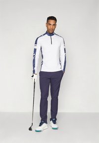 Camisa de golfe branca com fecho de correr no pescoço e mangas com padrão azul-marinho, combinada com calças azul-marinho e sapatos de golfe brancos com detalhes em verde-azulado. Taco de golfe segurado na mão esquerda.