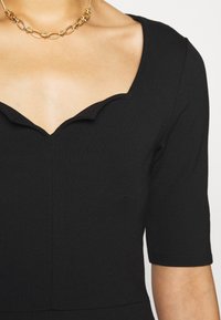 Zwarte jurk met een ronde halslijn en een scalloped rand. Korte mouwen, aansluitende textuur en subtiele naaddetails. Gouden ketting als accent.