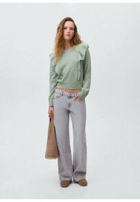Svetlozelen pulover z nagubanimi detajli, v kombinaciji s svetlo sivo širokima hlačami in rjavimi natikači. Manekenka drži rjavo torbico.