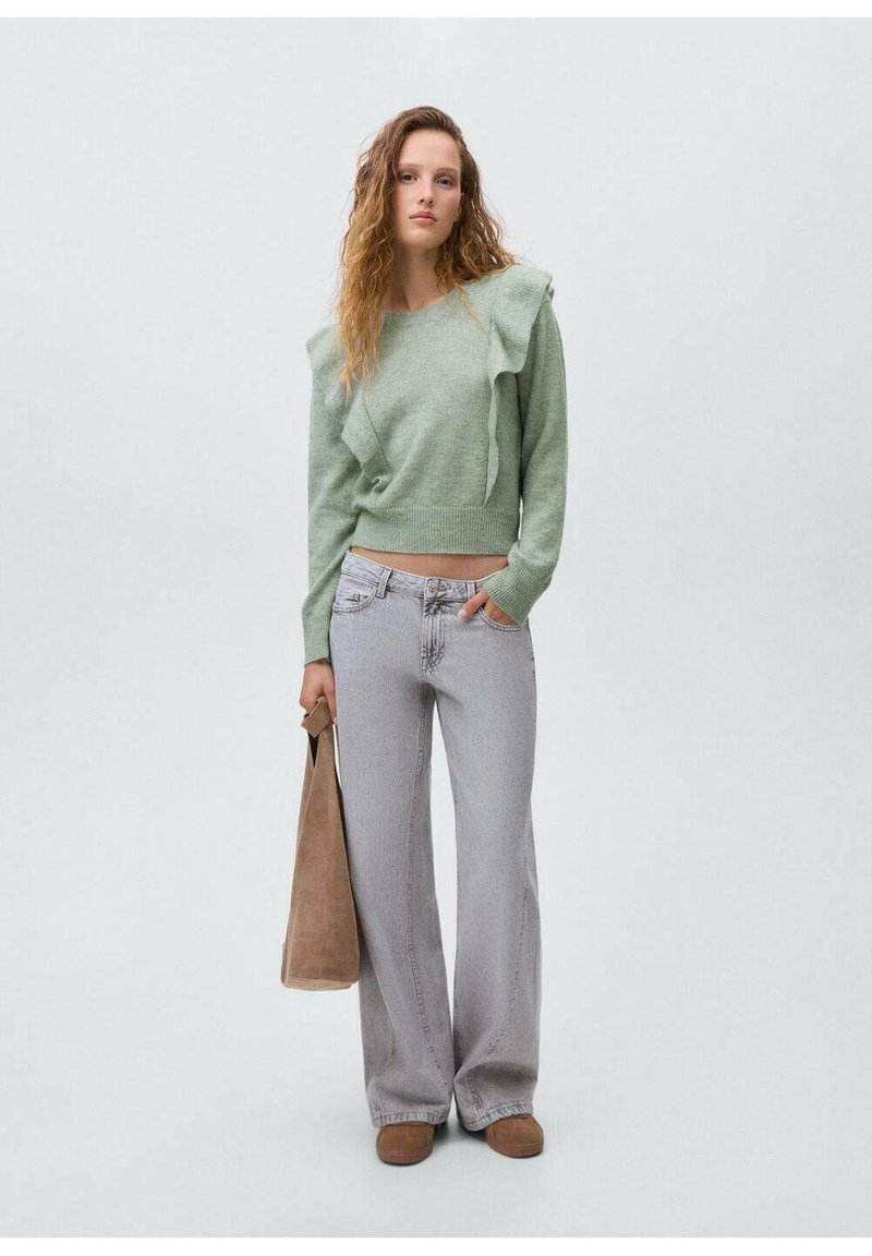 Svetlozelen pulover z nagubanimi detajli, v kombinaciji s svetlo sivo širokima hlačami in rjavimi natikači. Manekenka drži rjavo torbico.