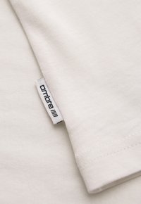 Gros plan sur un tissu blanc avec une petite étiquette blanche cousue dans la couture montrant le nom de marque noir « ombre » et le logo.