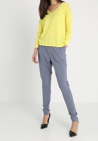 Donna con un maglione giallo a V, pantaloni grigi a sigaretta con tasche e scarpe con tacco nere, in piedi su uno sfondo bianco.