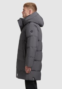 Sötétszürke párnázott parka magas gallérral és levehető kapucnival, kerek logóval ellátott folttal és oldalsó zsebekkel. Simább textúrájú és közép combig érő hosszúságú.