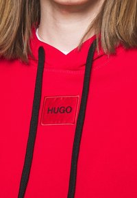 Czerwona bluza z kapturem wykonana z bawełny, z kapturem na sznurki, czarnymi sznurkami i prostokątną naszywką z nazwą marki "HUGO" w czarnym tekście.
