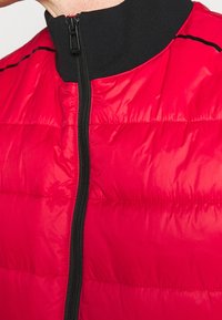 Primo piano di una giacca trapuntata rossa con cerniera nera e colletto alto indossata sulla parte superiore del petto e del collo di una persona.