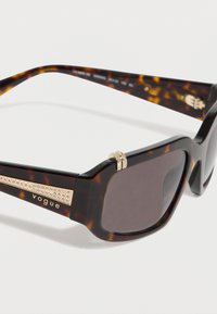 Γυαλιά ηλίου καφέ με ορθογώνια σκούρα φακούς, διακοσμήσεις σε χρυσό τόνο και λεπτομέρεια με στρας στη γέφυρα των γυαλιών, με το λογότυπο "vogue".