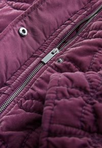 Gesteppter violetter Stoff mit glatter Textur, versehen mit einem silbernen Reißverschluss und Druckknopfverschluss, der die detaillierte Nahtarbeit betont.