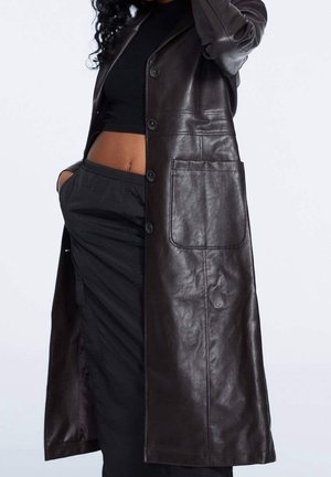 Femme portant un long manteau en cuir marron foncé par-dessus un crop top noir et un pantalon, les mains dans les poches du pantalon, sur un fond clair uni.