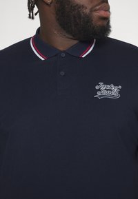 Marinblå polo skjorta med tvåknäppning, detaljer i röd och vit randig krage, och en vit broderad logotyp på bröstet.