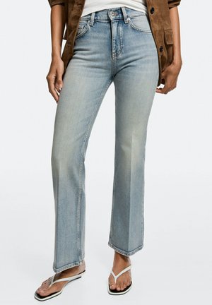 Personne portant un jean droit bleu clair, des sandales tongs blanches, une chemise blanche et une veste marron aux manches retroussées.