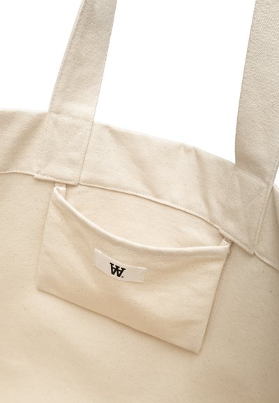 Sac en toile de couleur crème, avec une poche extérieure unique portant une étiquette. Deux poignées en tissu attachées en haut.