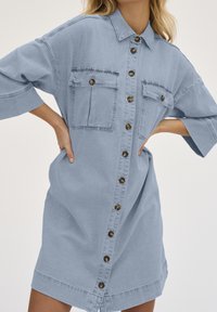 Robe chemise en denim bleu clair avec des manches courtes, fermeture boutonnée sur le devant, deux poches poitrine et une coupe décontractée. Elle présente des boutons en bronze et un ourlet arrondi.