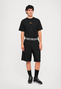 Νεαρός άνδρας στέκεται, φορώντας μαύρο μπλουζάκι, μαύρες βερμούδες με λευκή ζώνη Versace Jeans Couture, μαύρες κάλτσες και μαύρα αθλητικά παπούτσια.