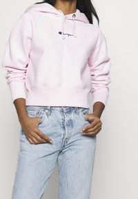 Sudadera con capucha de color rosa claro con cordones blancos y logotipo bordado; combinada con jeans de tiro alto y azul deslavado.