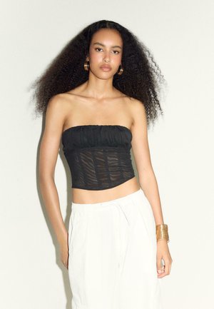 Femme aux cheveux bouclés et volumineux portant de grandes boucles d’oreilles créoles, un top noir sans bretelles en mesh, un pantalon blanc et un bracelet manchette doré, debout contre un mur blanc.