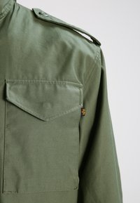 Cămașă de stil militar de culoare kaki, cu epaulet butonat pe umăr, un singur buzunar pe piept și un mic etichetă de marcă lângă buzunar.