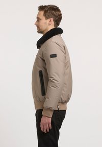 Veste bomber brun clair avec un col en fausse fourrure noir, poche zippée sur le côté, poignets et ourlet côtelés. Matériau lisse et rembourré.