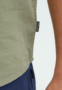 Chemise en coton vert olive avec un ourlet arrondi, dotée d'une étiquette noire sur le côté. Le tissu présente une apparence douce et texturée.