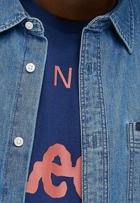 Close-up van een persoon die een blauw shirt met gedeeltelijk zichtbare rode tekst draagt onder een ongebottonde blauwe spijkerjack.