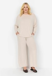 Set da salotto beige chiaro composto da un top a maniche lunghe con vestibilità comoda e pantaloni a gamba larga. Tessuto morbido con tasche sui lati. Scarpe nere senza lacci.