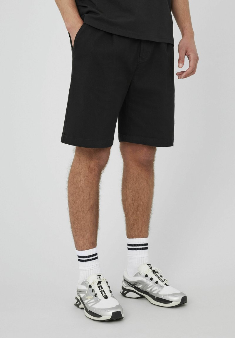 Shorts noirs en coton avec poches latérales, présentant une coupe droite et un ourlet. Associés à des chaussures de sport blanches et à des chaussettes rayées blanches et noires.