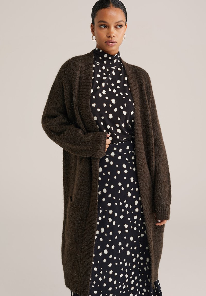 Smeškan smeđi pleteni kardigan s otvorenim prednjim dijelom, s dva džepa i rebrastim manžetama, nosi se preko tamne haljine s bijelim točkicama.