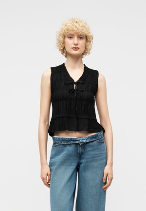 Kvinne med krøllete blondt hår har på seg en ermeløs svart bluse med rysjer og knytedetaljer, samt løstsittende blå høytlivsjeans med brettet midjebånd.