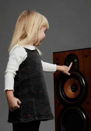 Barn i en mörk denimklänning med blommönster, som rör vid en trähögtalare med en stor cirkulär högtalarkon och svarta accenter.